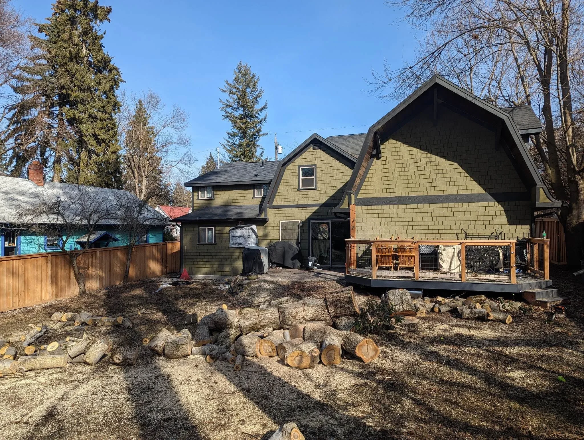 Residential backyard with stacked logs from tree removal, wooden deck, and fencing visible